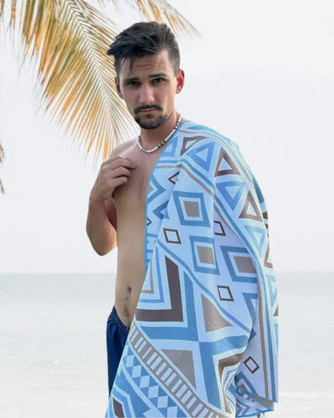 Man standing by the sea, wearing swim pants and looking at the camera, with an azure-patterned towel draped over his shoulder. Neutral-toned sea and palm leaf in the background.