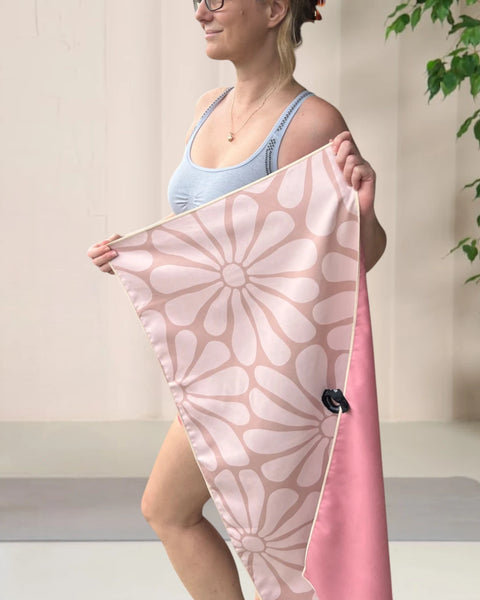 Woman standing in a yoga room, wearing a yoga outfit, facing sideways and holding a beige coloured gym towel with a floral pattern, stretched out with both hands.