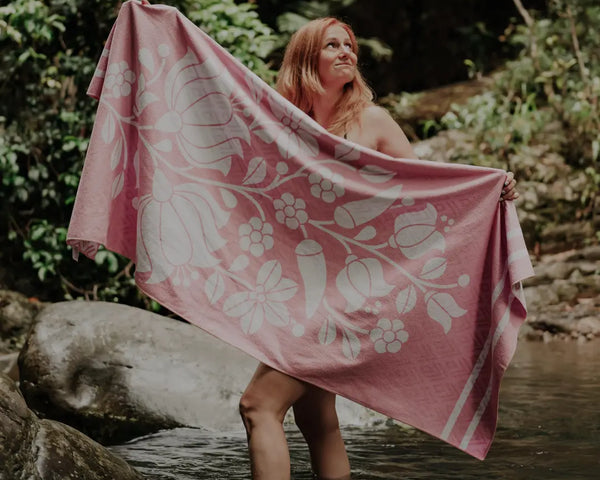 Woman standing in a forest lake, holding a pink towel stretched out in front of her.