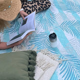 Woman lying on a light green palm leaf-patterned picnic blanket, reading a book.