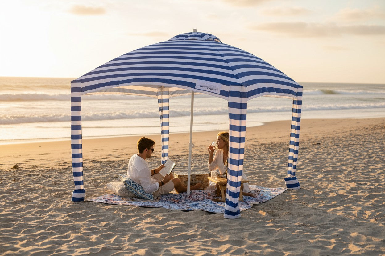 Two people sitting under a blue striped beach umbrella on a mandala picnic mat on a sandy beach.