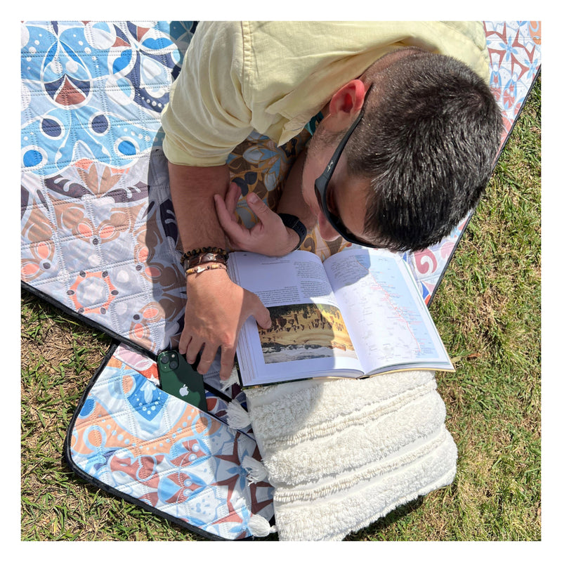 A man is lying on a mandala pattern picnic blanket with a zip pocket, reading and securing his phone into the pocket.