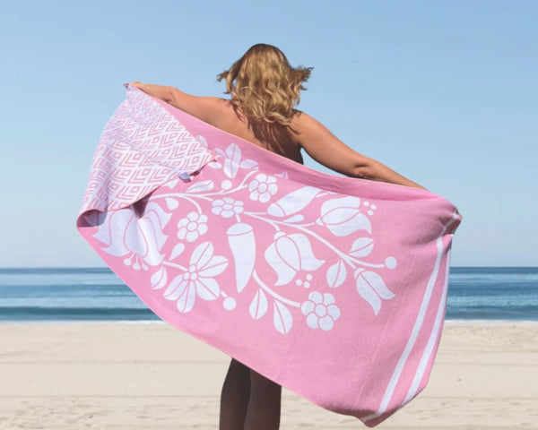 Woman standing on the beach with her back to the camera, holding a pink towel stretched out behind her.