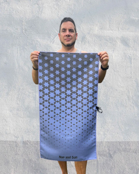 Man standing in front of a wall, facing the camera, holding an indigo-coloured gym towel with a black star pattern, stretched out with both hands.