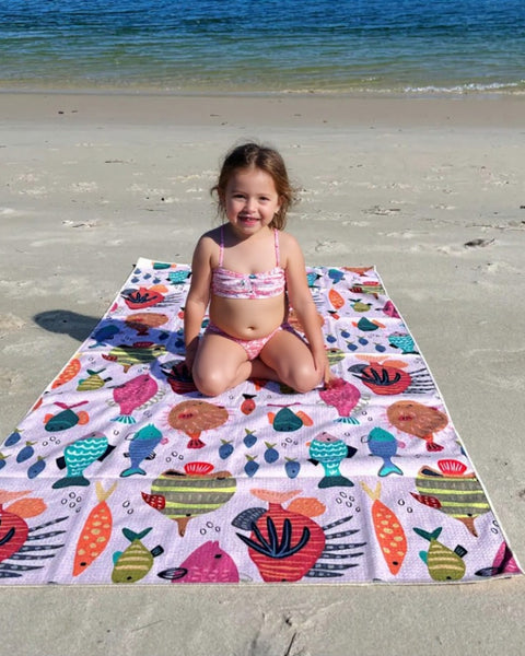 Child sitting on a colorful beach towel with a sea-themed pattern on a sandy beach.