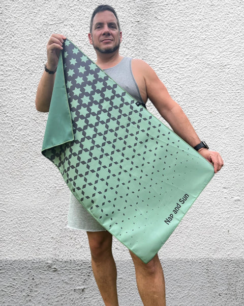 Man standing in front of a wall, facing the camera, holding a green-coloured gym towel with a black star pattern, stretched out with both hands.