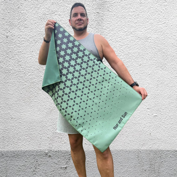 Man standing in front of a wall, facing the camera, holding a green-coloured gym towel with a black star pattern, stretched out with both hands.