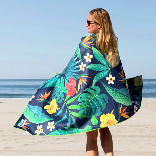Woman standing on the beach with her back to the camera, wrapped in a Tropical Bliss towel featuring green tropical leaves and hibiscus flowers.