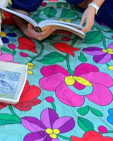 Close up of a woman lying on her side on a vibrant, flower-patterned picnic blanket, reading a book in a peaceful park setting.