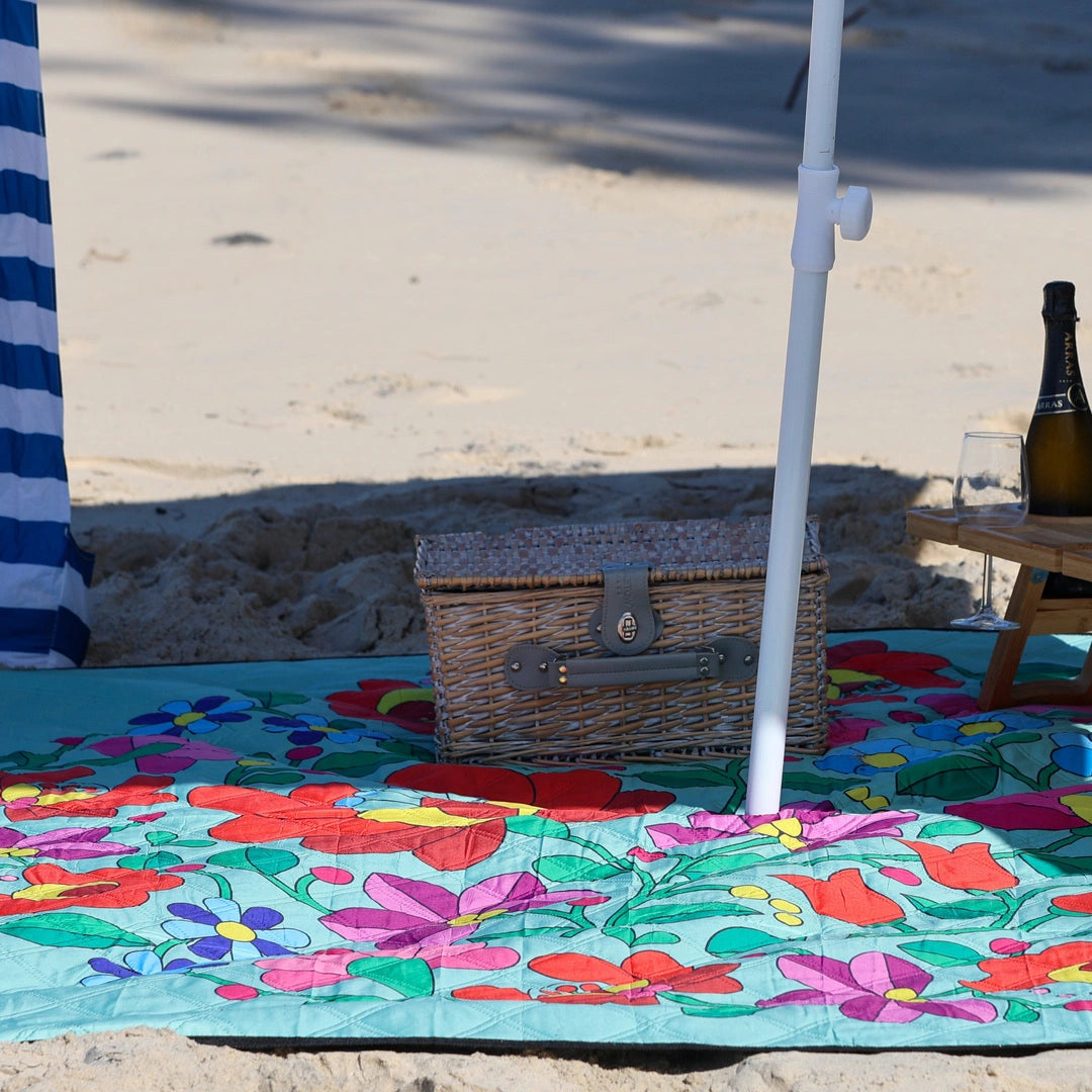Close-up of Nap and Sun brand floral pattern waterproof picnic blanket on the beach with cabana attached and picnic basket placed on top.