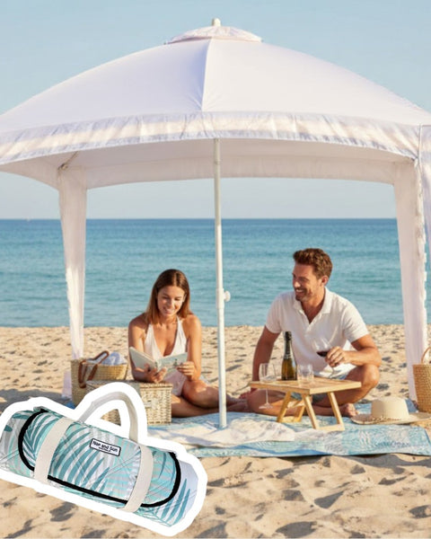 Leafy picnic blanket set up on a sandy beach under a cabana, with people relaxing and enjoying a picnic.