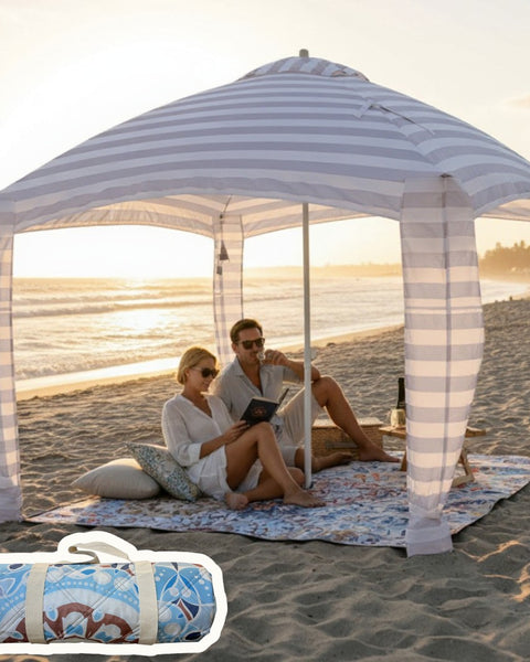Two people sitting under a cabana on a mandala patterned picnic blanket at the beach.