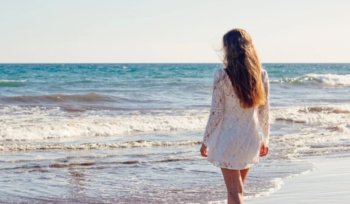 Woman in a short white dress seen from behind, stepping barefoot into the sea on a sandy beach.