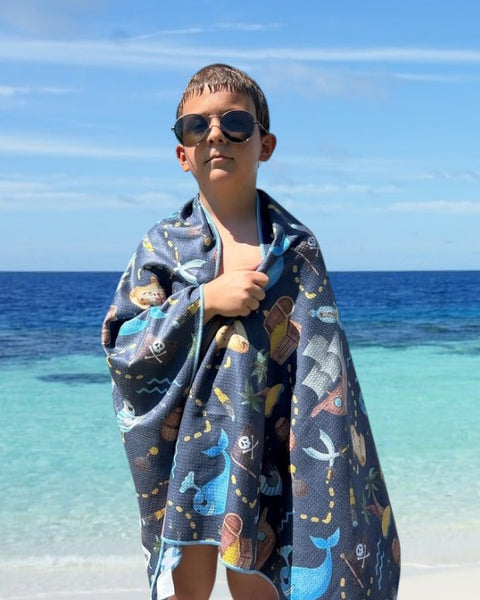 A child stands by a beach, wrapped snugly in a colorful pirate theme beach towel. Towel name: Napandsun Treasure Island sand-free beach towel for kids.