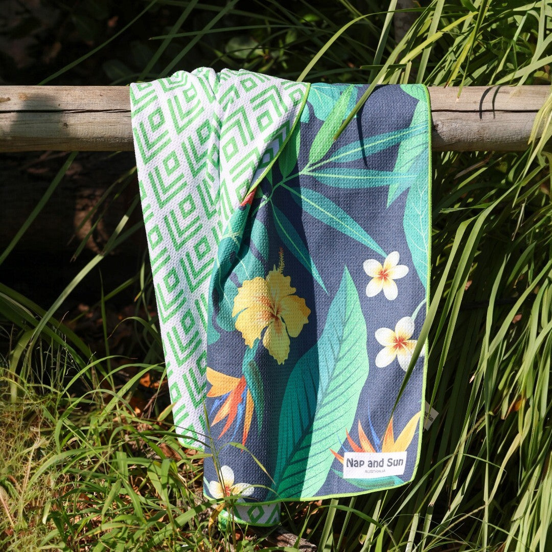 Nap and Sun brand tropical-pattern sand-free beach towel hanging on a wooden fence at the beach, partially framed by coastal bush.