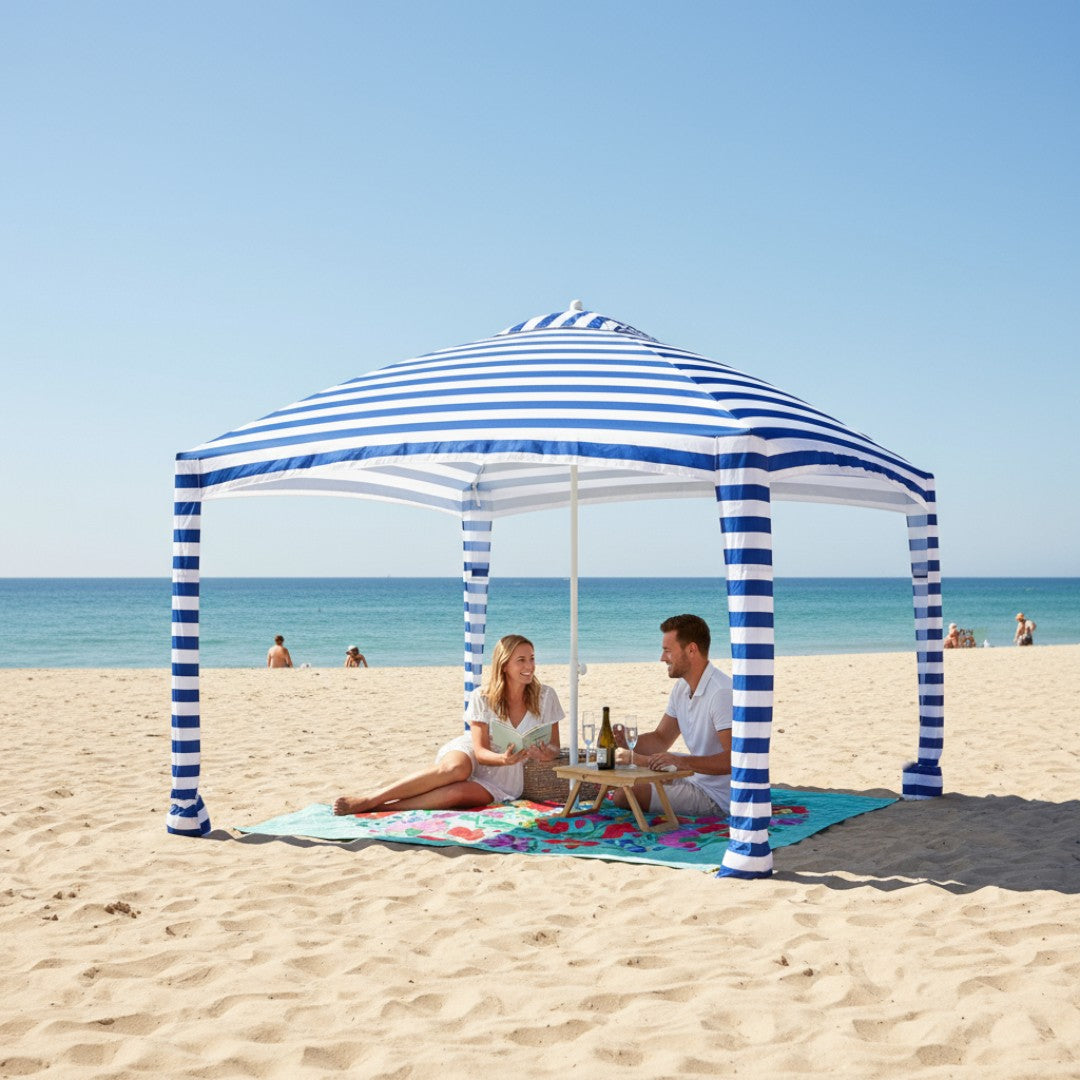Two people sitting under a striped canopy on a colourful picnic blanket on a sandy beach