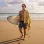 man with a surfboard and a yellow beach towel on his shoulder is walking on the beach