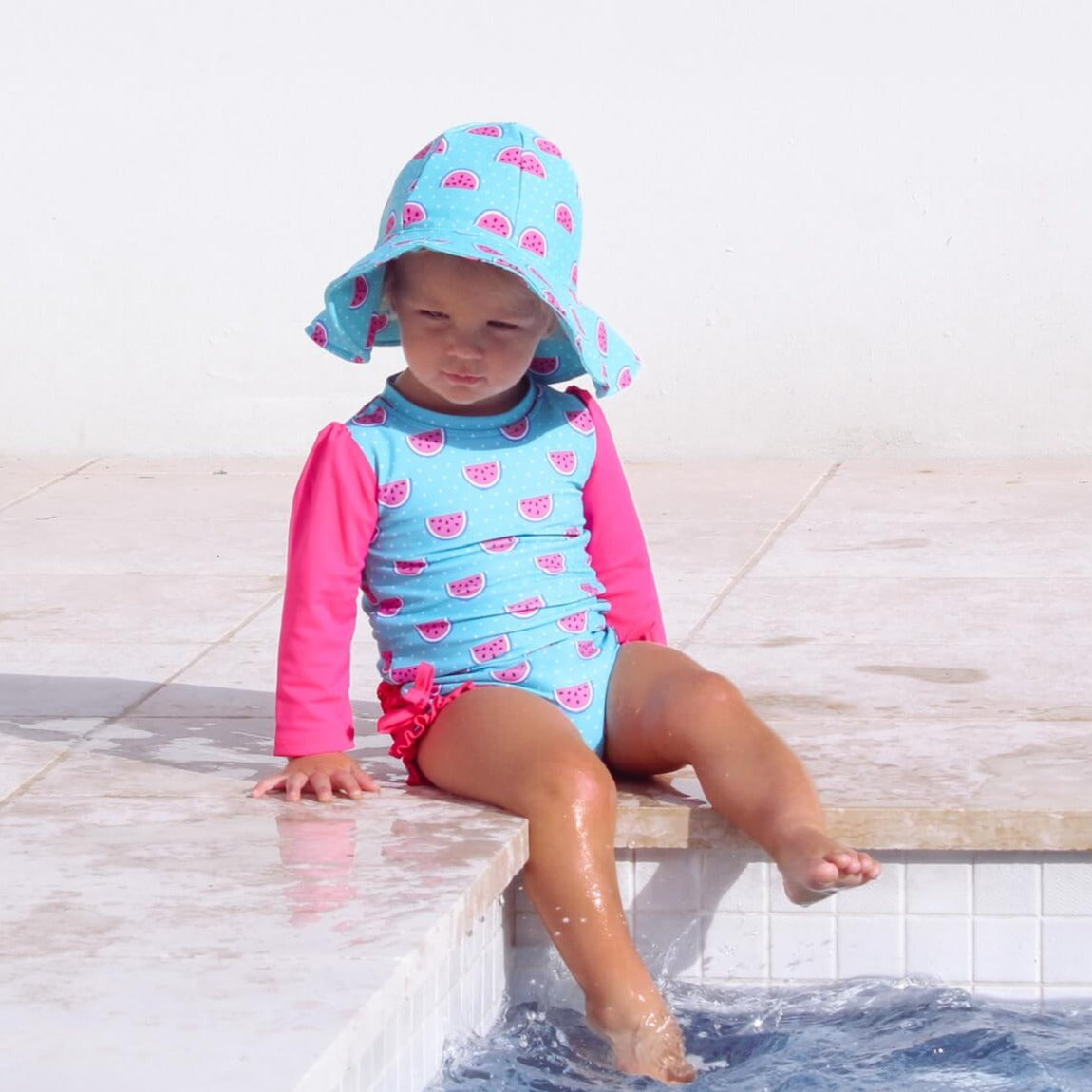 Child wearing a blue and pink swimsuit with a matching hat, sitting by a pool.