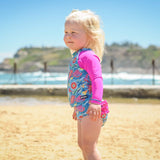 Child in a colorful swimsuit with pink sleeves standing on a beach.