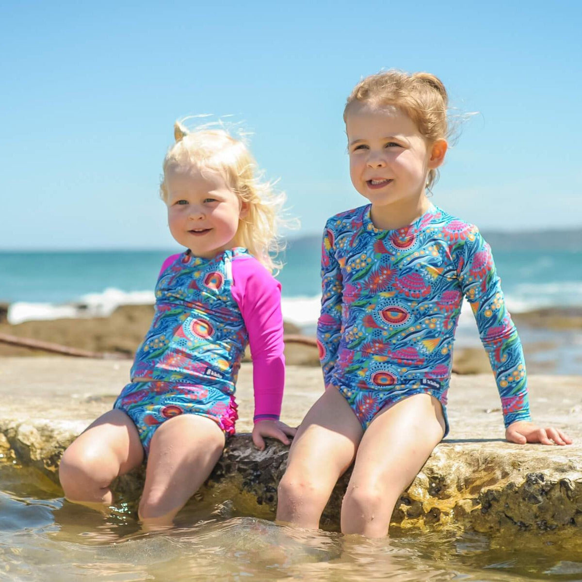 Two children in colorful swimsuits sitting on a rock by the ocean.