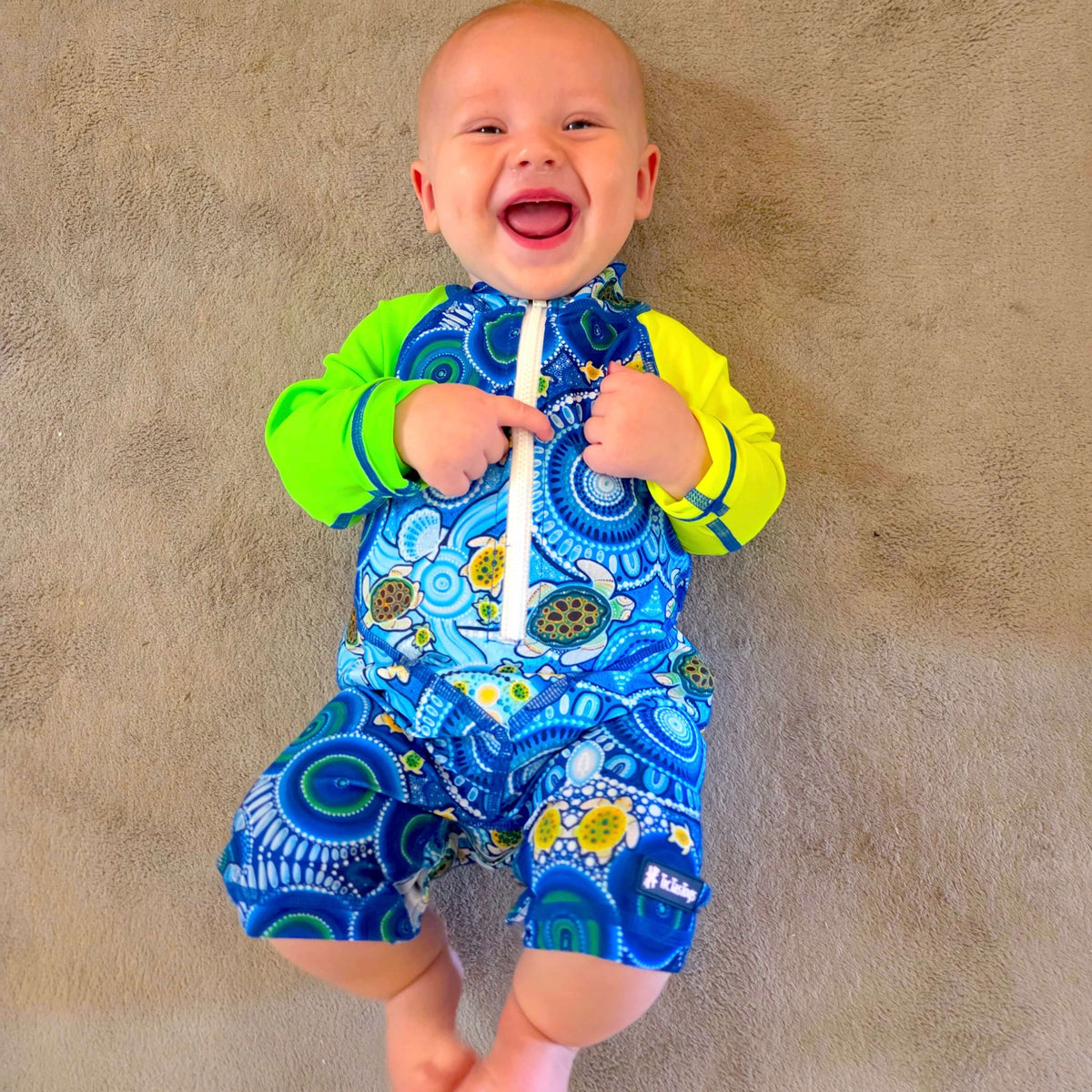 Baby wearing a blue and green swimsuit with turtle design on a beige carpet