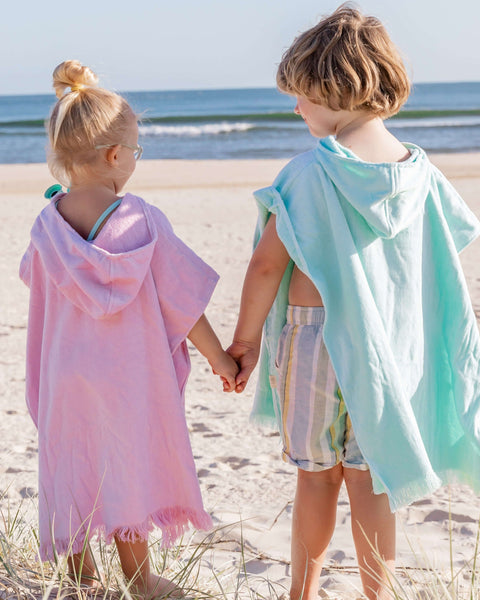 Two children holding hands on a beach, wearing colorful hooded beach towels.