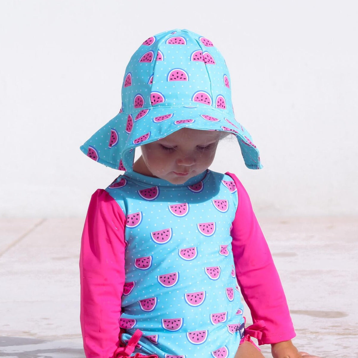 Child wearing a blue and pink watermelon-patterned swimsuit and hat on a light wooden floor.