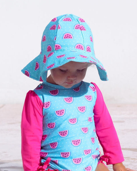 Child wearing a blue and pink watermelon-patterned swimsuit and hat on a light wooden floor.