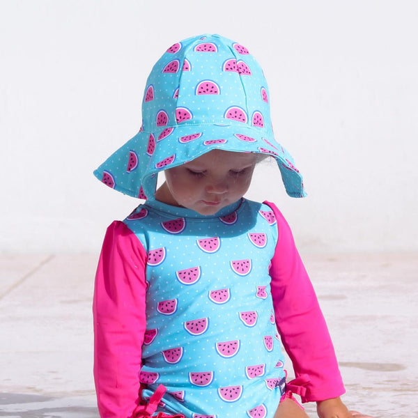 Child wearing a blue and pink watermelon-patterned swimsuit and hat on a light wooden floor.