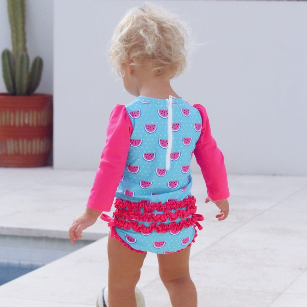 Child wearing a blue and pink swimsuit with watermelon pattern on a white surface.