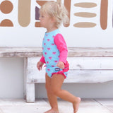 Child wearing a blue and pink swimsuit with heart patterns, standing on a wooden floor.