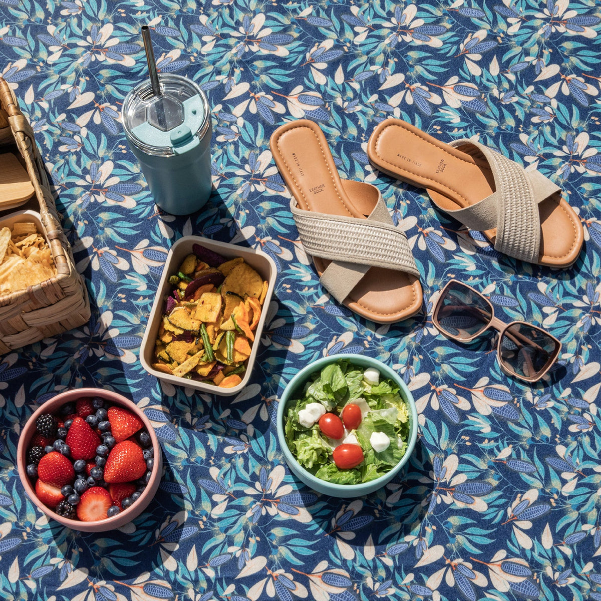 Outdoor setting with sandals, salad, snacks, and a drink on a blue floral tablecloth.