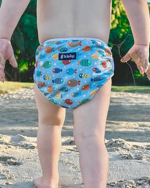 Child wearing a colorful fish-patterned swim diaper on a sandy beach.