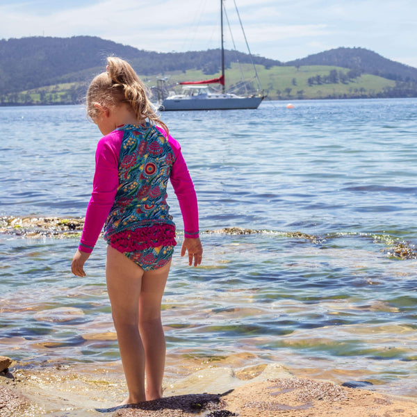 Child in a colorful swimsuit standing on a rocky beach with a sailboat in the background.