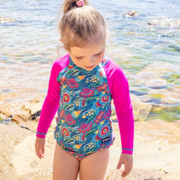 Child wearing a colorful swimsuit with pink sleeves standing on a beach.