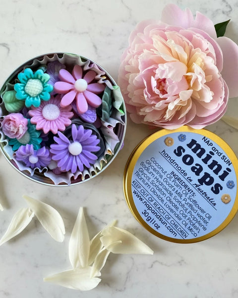 A round gold tin labeled 'mini soaps' with several colorful flower-shaped handmade soaps arranged on a white marble surface. The tin is open and there is a pink peony next to the tin.