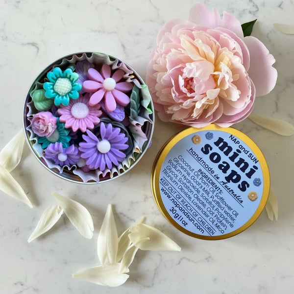 A round gold tin labeled 'mini soaps' with several colorful flower-shaped handmade soaps arranged on a white marble surface. The tin is open and there is a pink peony next to the tin.