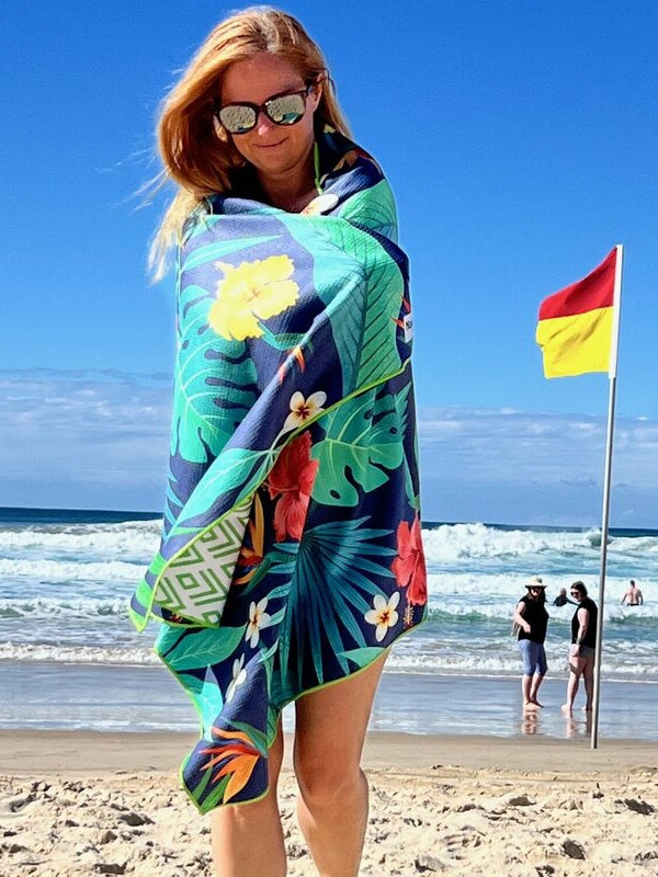 A woman wrapped in a tropical beach towel, standing on a sandy beach with the ocean in the background.