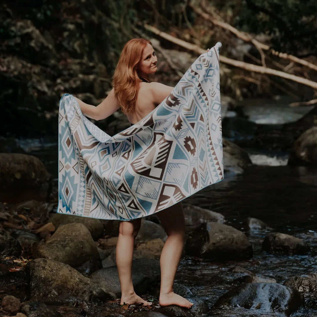 Back view of a woman standing in a forest in front of a waterfall, holding a light blue, brown, and white tribal-style towel stretched out behind her. The towel called: Azure Tribe from Nap and Sun