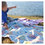 A toddler sits on a colorful beach towel by the lake, watching intently and pointing into the distance, surrounded by the serene natural beauty of the water. Napandsun Rainbow Reef sand free towel for Kids, Free wetbag included