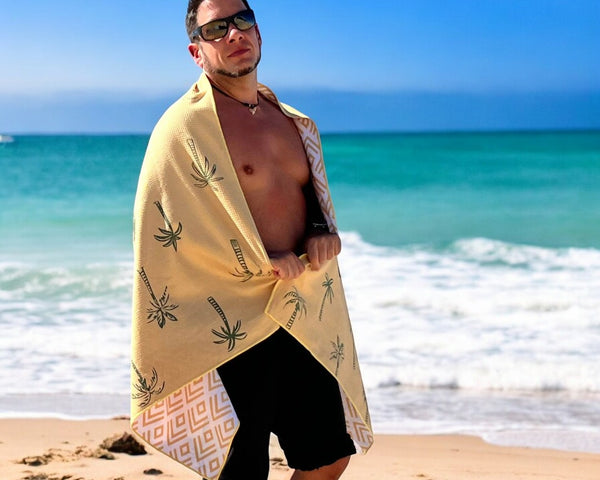 Man stands on soft sand next to the sea, facing the camera. He is wrapped in a yellow-gold Nap and Sun Sunset Palm beach towel.