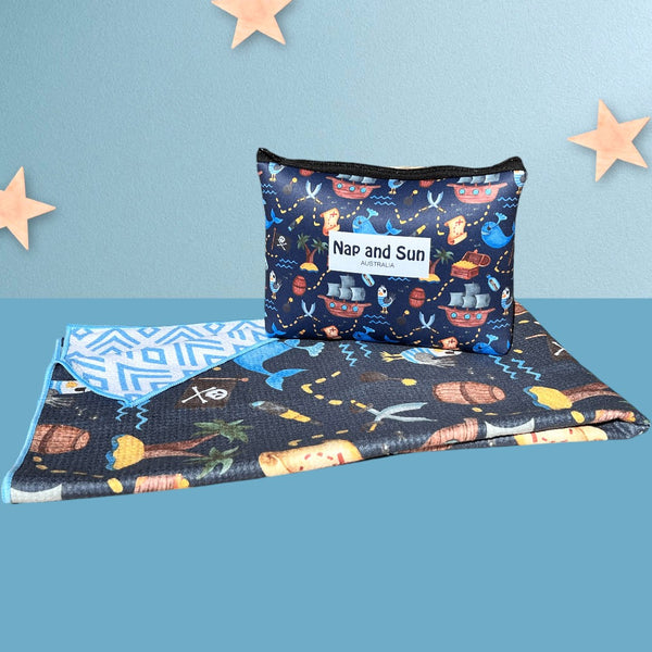 Napandsun Treasure Island sand-free beach towel for kids featuring a double sided design. Front: Pirate-themed design with whales, islands, treasure map, and pirate ships on a dark blue background. Back: white background with blue geometric lines. Free matching wet bag is included. 