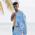 Man standing by the sea, wearing swim pants and looking at the camera, with an azure-patterned towel draped over his shoulder. Neutral-toned sea and palm leaf in the background.