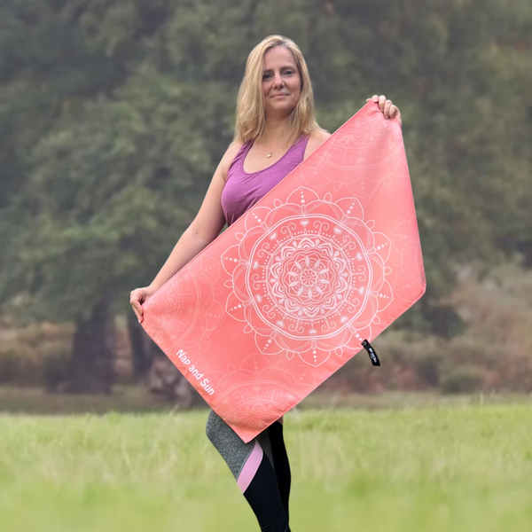 Woman standing in a park, wearing a yoga outfit, facing the camera and holding a pink mandala-patterned gym towel stretched out with both hands.
