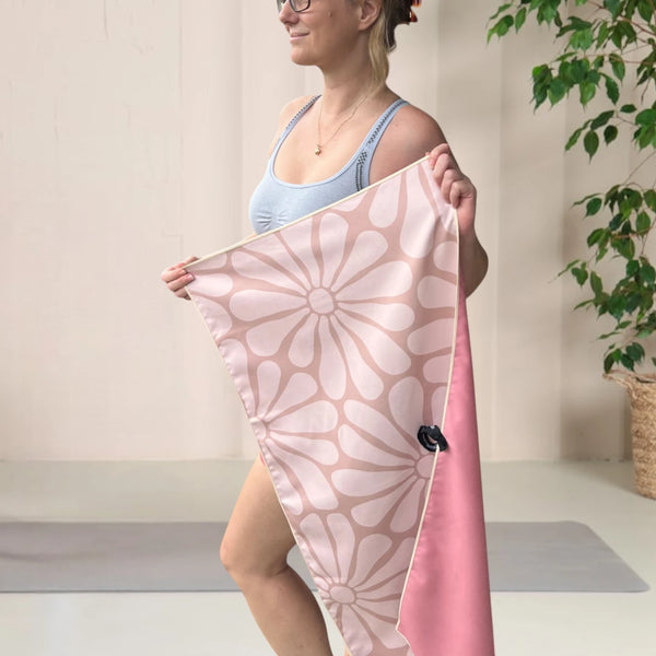 Woman standing in a yoga room, wearing a yoga outfit, facing sideways and holding a beige coloured gym towel with a floral pattern, stretched out with both hands.
