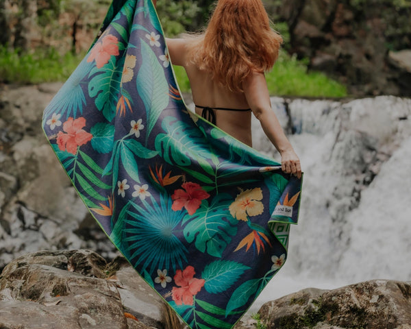 Woman standing in front of a waterfall, holding a tropical pattern towel stretched behind her.