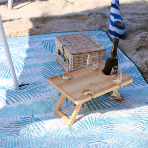 Close-up of a light green palm-leaf print picnic blanket on the beach, set up with a cabana, picnic basket, champagne bottle, and glasses on the blanket.