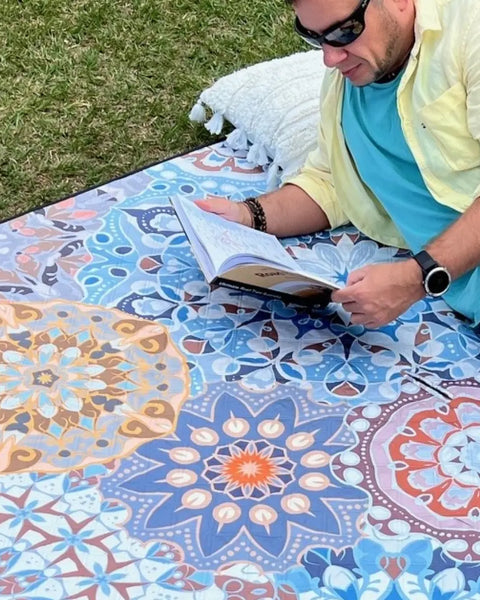 Close-up of a man lying on a mandala-patterned picnic blanket in a park, reading a book.