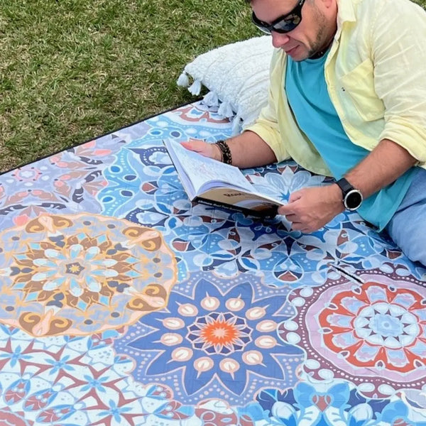Close-up of a man lying on a mandala-patterned picnic blanket in a park, reading a book.