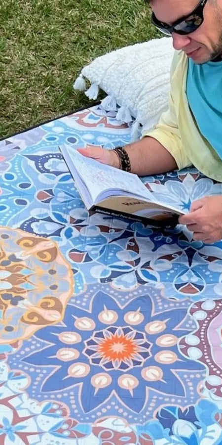 Close-up of a man lying on a mandala-patterned picnic blanket in a park, reading a book.